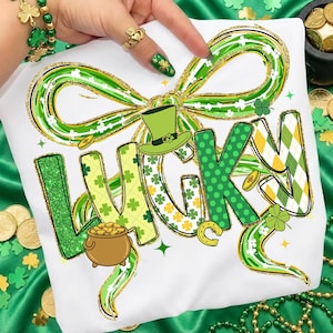 May include: White t-shirt with a St. Patrick's Day design. The word "LUCKY" is displayed in green, yellow, and white, with a green bow and a pot of gold. The design includes shamrocks, a horseshoe, and a leprechaun hat.