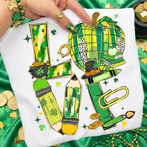 May include: White t-shirt with a colorful graphic design featuring the word "LOVE" composed of St. Patrick's Day themed elements. Includes a pot of gold, pencils, an apple, and a ruler. The design incorporates green, yellow, and gold accents.