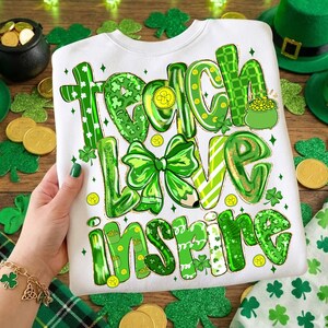 May include: White sweatshirt featuring the words "Teach Love Inspire" in green, adorned with shamrocks, a bow, and a pot of gold. Ideal for St. Patrick's Day or as a gift for a teacher.
