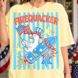 Pode incluir: Camiseta amarela com listras verticais azuis e a palavra "FIREQUACKER" em vermelho. A camiseta apresenta um pato de desenho animado usando um chapéu patriótico e segurando uma bandeira americana, montando um foguete azul com gráficos de estrelas e fogos de artifício.