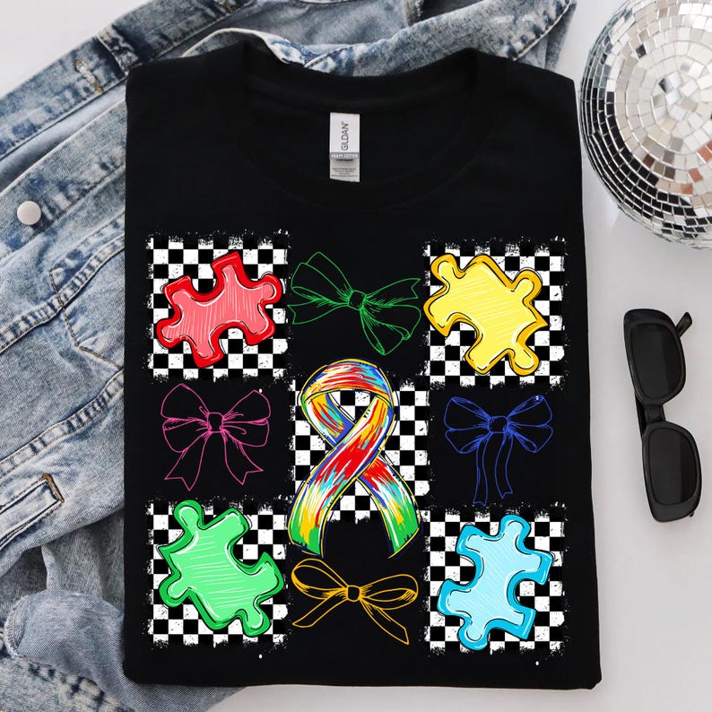May include: Black t-shirt with a design of colourful puzzle pieces and ribbons on a chequered background. A tie-dye ribbon is in the centre. The shirt is folded on a denim jacket.