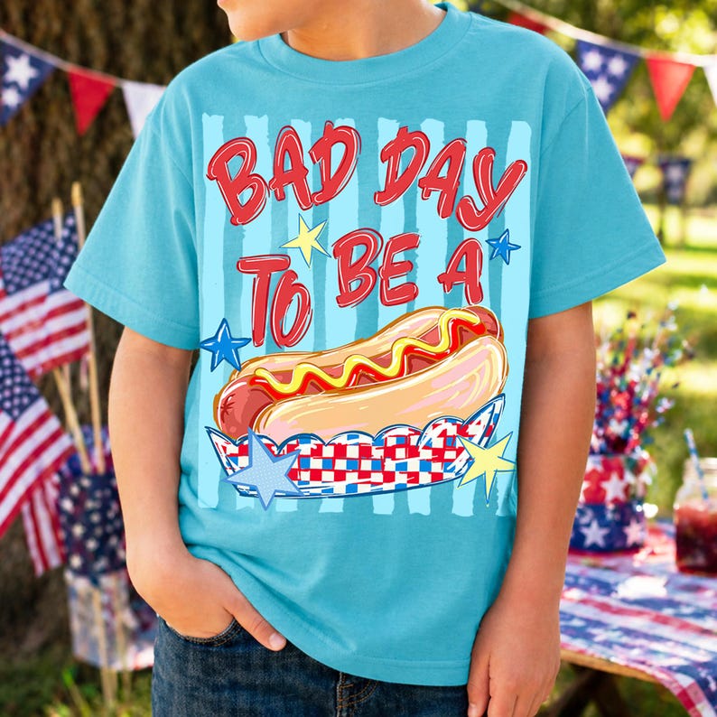 以下が含まれることがあります： 「BAD DAY TO BE A」の赤い文字と漫画のホットドッグのグラフィックがプリントされた水色のTシャツ。 ホットドッグは黄色のマスタードで、星と赤、白、青の模様が入った装飾的なホルダーに入っています。