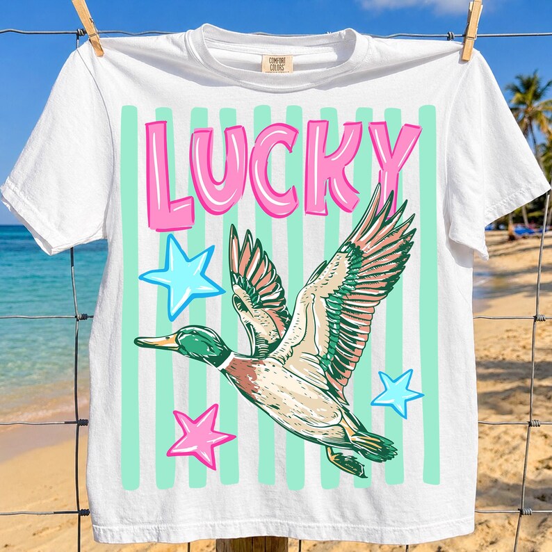 Puede incluir: Camiseta blanca con la palabra "LUCKY" en letras rosas con burbujas. Debajo, un gr&aacute;fico de un pato volando, en tonos verdes, marrones y rosas, acompa&ntilde;ado de estrellas azules y rosas y rayas verticales verde menta.