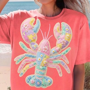 Könnte beinhalten: Korallfarbenes T-Shirt mit einem Patchwork-Hummer-Design. Der Hummer ist mit Blumenmustern in Pastellfarben verziert, die mit weißen Perlen umrandet sind. Das T-Shirt hat kurze Ärmel und einen Rundhalsausschnitt, perfekt für Freizeitkleidung.