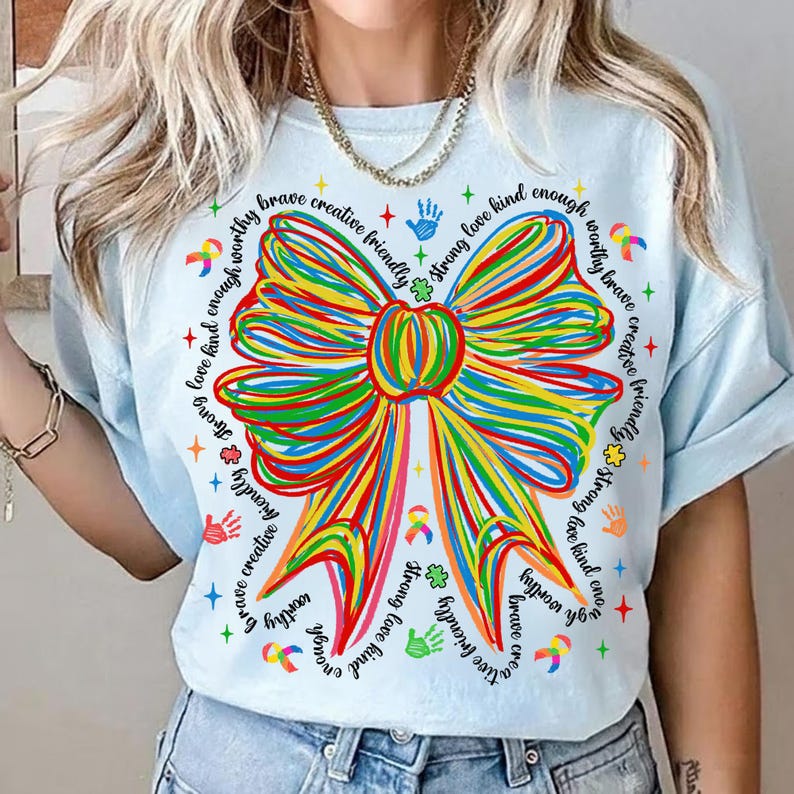 May include: Light blue t-shirt featuring a large, colorful bow design. The bow is made of multiple lines in various colors. The shirt also has text around the bow that says "brave, creative, friendly, strong, love, kind, enough, worthy".