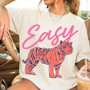 May include: Cream-colored t-shirt with the word "Easy" in pink script above a coral-colored tiger with navy blue stripes. The shirt is a casual, oversized style.