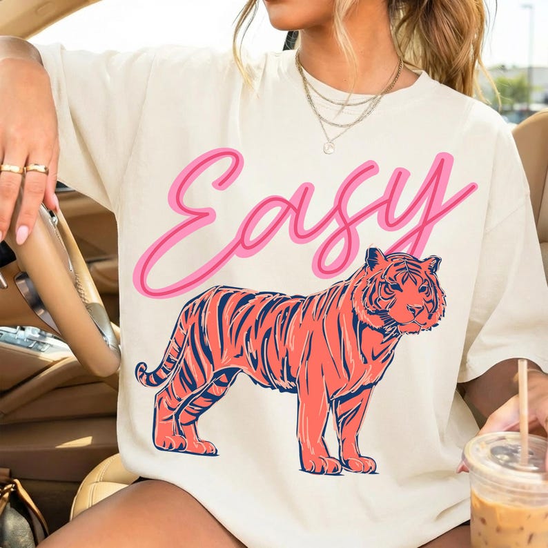May include: Cream-colored t-shirt with the word "Easy" in pink script above a coral-colored tiger with navy blue stripes. The shirt is a casual, oversized style.