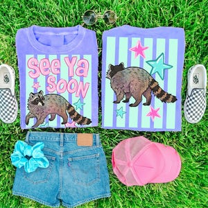 May include: Two purple t-shirts with a raccoon graphic and the text "See Ya Soon". The image also includes denim shorts, a pink trucker hat, a blue scrunchie, and checkered slip-on sneakers, all on a green grassy surface.