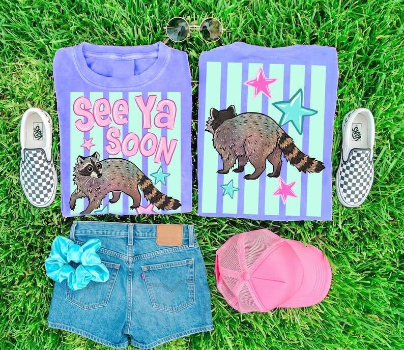 May include: Two purple t-shirts with a raccoon graphic and the text "See Ya Soon". The image also includes denim shorts, a pink trucker hat, a blue scrunchie, and checkered slip-on sneakers, all on a green grassy surface.