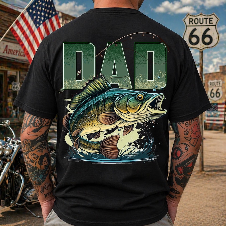 Pu&ograve; includere: T-shirt nera con un'illustrazione verde e blu di un pesce che salta fuori dall'acqua, con la parola "DAD" in lettere verdi sopra. Il design include una lenza. Lo sfondo mostra un cartello della Route 66 e una bandiera americana.