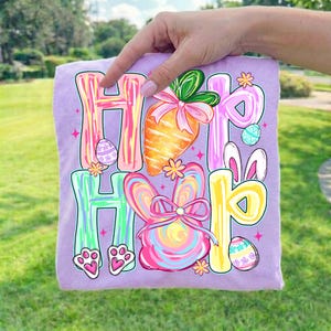 Peut inclure: Un t-shirt lilas avec un motif de P&acirc;ques color&eacute;. Le graphisme comprend les mots "Hip Hop" en grandes lettres stylis&eacute;es, ainsi qu'une carotte, un lapin et des &oelig;ufs de P&acirc;ques. Le t-shirt est tenu par une main.