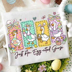 May include: White t-shirt with a colorful Easter-themed graphic. The design features four cartoon geese dressed in Easter attire, each within a different colored frame. The text "Just An Egg-cited Goose" is written below the geese.