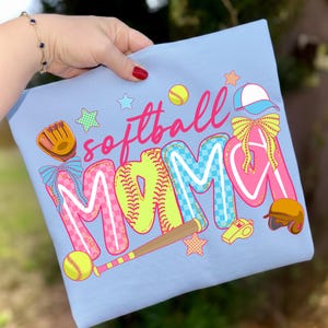 May include: Light blue t-shirt with the words "softball MAMA" in a colorful design. The design includes baseballs, a glove, a bat, a helmet, a whistle, and stars. The letters are outlined in white and filled with pink, blue, and yellow.