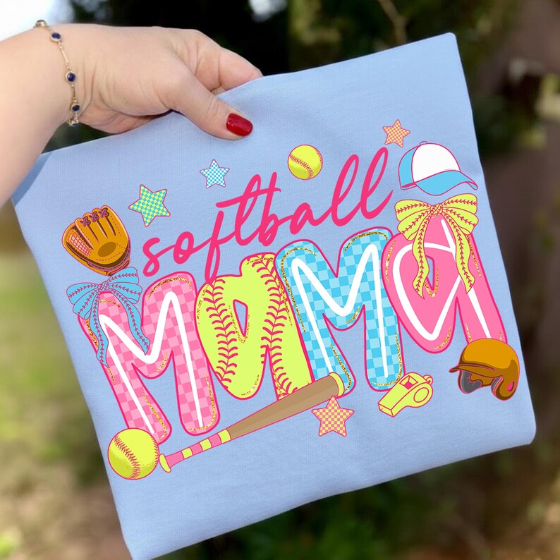 May include: Light blue t-shirt with the words "softball MAMA" in a colorful design. The design includes baseballs, a glove, a bat, a helmet, a whistle, and stars. The letters are outlined in white and filled with pink, blue, and yellow.
