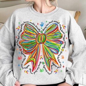 May include: A light gray sweatshirt featuring a large, colorful bow design. The bow is made of rainbow-colored lines. The words "brave", "creative", "friendly", "strong", "love", "kind", and "worthy" are written around the bow.