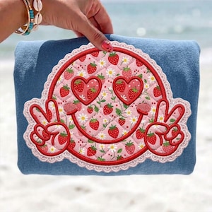 Könnte beinhalten: Eine blaue Denim-Tragetasche mit einem lächelnden Gesichtsdesign. Das Gesicht hat herzförmige Augen und Friedenszeichen, gefüllt mit Erdbeeren und Gänseblümchen und rot umrandet mit weißer Spitzenborte. Die Tasche wird von einer Person gehalten.