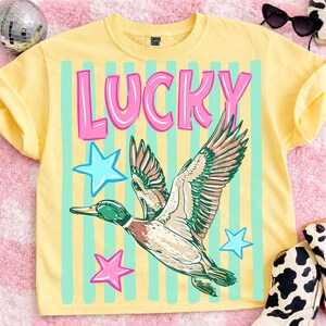 Puede incluir: Camiseta amarilla con la palabra "LUCKY" en letras rosas, un gr&aacute;fico de pato volando y estrellas azules y rosas. El pato tiene detalles verdes, marrones y blancos. Hay rayas celestes detr&aacute;s del gr&aacute;fico.