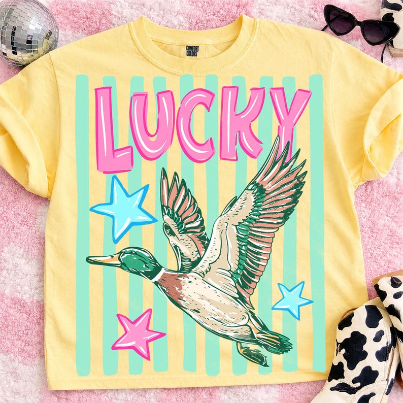 Puede incluir: Camiseta amarilla con la palabra "LUCKY" en letras rosas, un gr&aacute;fico de pato volando y estrellas azules y rosas. El pato tiene detalles verdes, marrones y blancos. Hay rayas celestes detr&aacute;s del gr&aacute;fico.