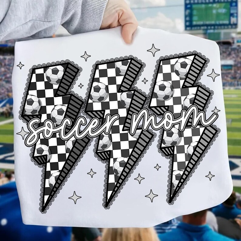 Puede incluir: Camiseta blanca con un dise&ntilde;o gr&aacute;fico de tres rayos. Cada rayo tiene un patr&oacute;n de cuadros blancos y negros con balones de f&uacute;tbol. Las palabras "soccer mom" est&aacute;n en cursiva. Estrellas y encaje adornan el dise&ntilde;o.