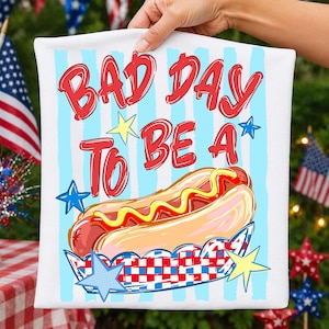 以下が含まれることがあります： 赤い文字で「BAD DAY TO BE A」と書かれた白いクッション。ホットドッグと星の漫画が描かれています。 ホットドッグは赤と青のチェック柄の紙トレイに入っており、青と黄色の星が付いています。