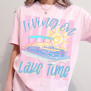 May include: Pink t-shirt with the words "Living on Lake Time" in blue script. The design features a blue and yellow pontoon boat with a sun graphic on a pink and white striped background.