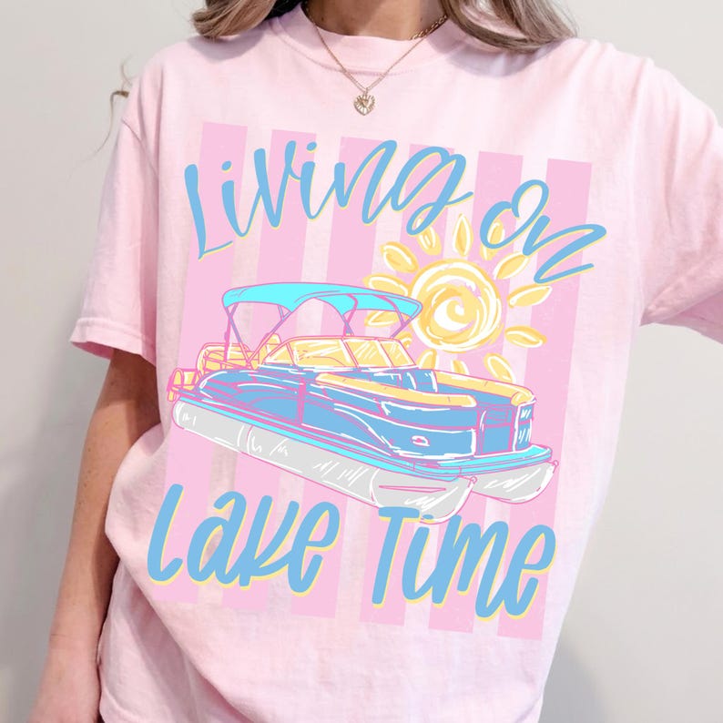 May include: Pink t-shirt with the words "Living on Lake Time" in blue script. The design features a blue and yellow pontoon boat with a sun graphic on a pink and white striped background.
