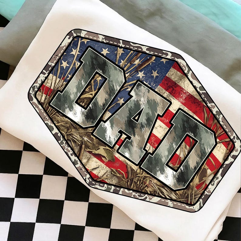 以下が含まれることがあります： 白いTシャツ。迷彩柄とアメリカ国旗のデザインで、メタリックな質感のフォントで「DAD」の文字が描かれています。デザインは迷彩の縁取りのある幾何学的な形の中にあります。Tシャツは白と黒のチェック柄の表面に折りたたまれています。
