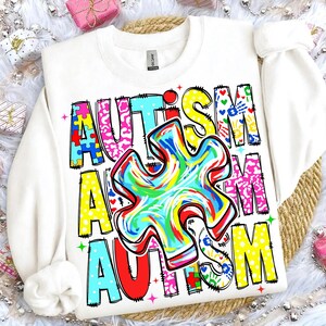 May include: White sweatshirt with the word "AUTISM" in colorful, block letters. The design includes a large, multi-colored puzzle piece in the center, with additional puzzle piece and handprint graphics. The sweatshirt has a crew neck and long sleeves.