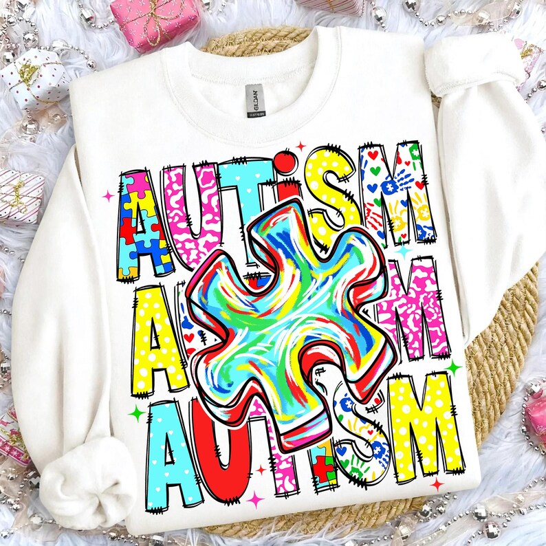 May include: White sweatshirt with the word "AUTISM" in colorful, block letters. The design includes a large, multi-colored puzzle piece in the center, with additional puzzle piece and handprint graphics. The sweatshirt has a crew neck and long sleeves.