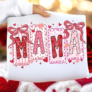 May include: White pillow with the word "MAMA" in a decorative font. The letters are in pink and red with heart and bow accents. The pillow is held by a person.