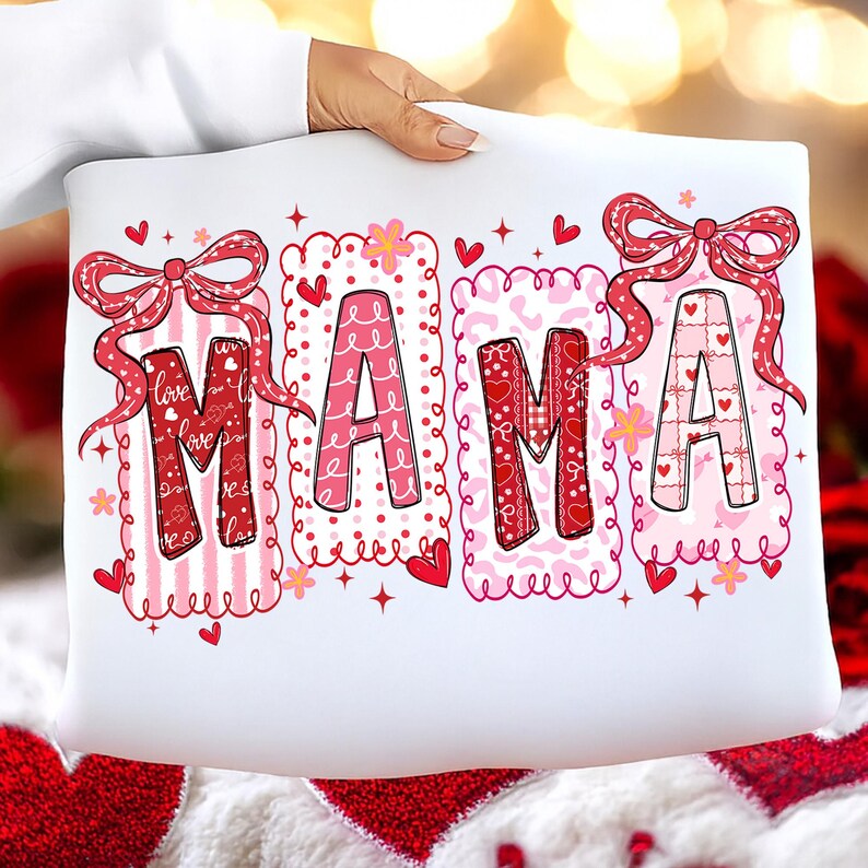 May include: White pillow with the word "MAMA" in a decorative font. The letters are in pink and red with heart and bow accents. The pillow is held by a person.