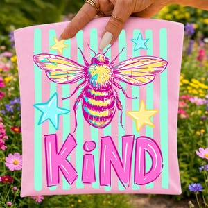 Pode incluir: Saco tote rosa com um desenho de abelha e estrelas. A abelha é amarela, rosa e azul, com a palavra "KIND" em letras rosa. O fundo tem listras verticais rosa e verde claro.
