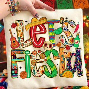 Könnte beinhalten: Ein cremefarbenes Kissen mit den Worten "Let's Fiesta" in farbenfrohen, festlichen Buchstaben. Das Design umfasst einen Sombrero, eine Gitarre, einen Totenkopf und andere mexikanische Illustrationen. Das Kissen wird von einer Person gehalten.