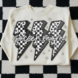 Puede incluir: Sudadera de color crema con un dise&ntilde;o gr&aacute;fico que presenta tres rayos con balones de f&uacute;tbol y un patr&oacute;n de cuadros. Las palabras "soccer mom" est&aacute;n escritas en una fuente cursiva. El dise&ntilde;o est&aacute; delineado con un ribete de encaje negro y formas de estrella.