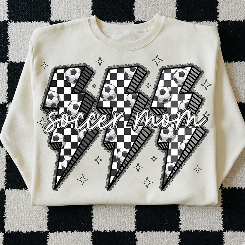 Puede incluir: Sudadera de color crema con un dise&ntilde;o gr&aacute;fico que presenta tres rayos con balones de f&uacute;tbol y un patr&oacute;n de cuadros. Las palabras "soccer mom" est&aacute;n escritas en una fuente cursiva. El dise&ntilde;o est&aacute; delineado con un ribete de encaje negro y formas de estrella.