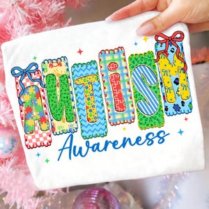 Puede incluir: Tela blanca con la palabra "AUTISMO" en letras coloridas y estampadas, cada una con un diseño único. Debajo, la palabra "Awareness" está escrita en azul. El diseño incluye piezas de rompecabezas, lazos y estrellas, promoviendo la concienciación sobre el autismo.