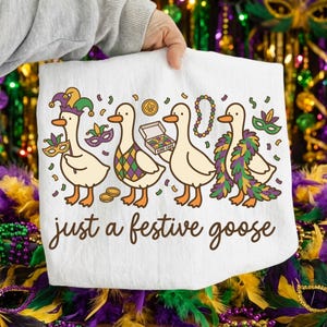Op de afbeelding: Witte decoratieve handdoek met vier cartoon ganzen gekleed voor Mardi Gras. De ganzen dragen feestelijke accessoires zoals maskers, kralen en een gevederde boa. De tekst "just a festive goose" is geschreven in een script.