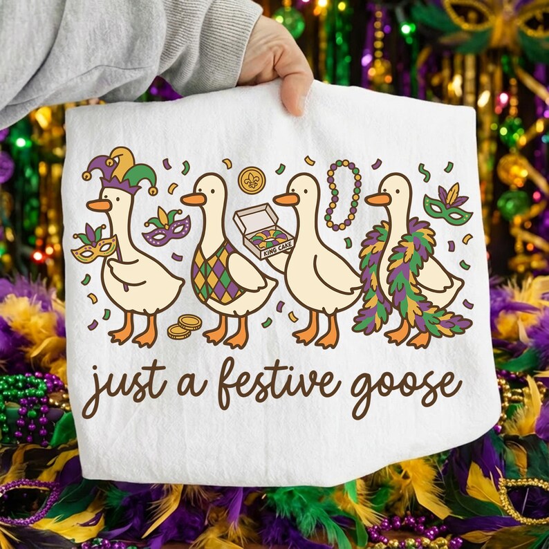 Op de afbeelding: Witte decoratieve handdoek met vier cartoon ganzen gekleed voor Mardi Gras. De ganzen dragen feestelijke accessoires zoals maskers, kralen en een gevederde boa. De tekst "just a festive goose" is geschreven in een script.