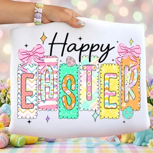 May include: White pillow with the words "Happy Easter" in black script. The word "EASTER" is in colorful, patterned blocks with pink bows and decorative elements. The pillow is held by a hand with a bunny bracelet.