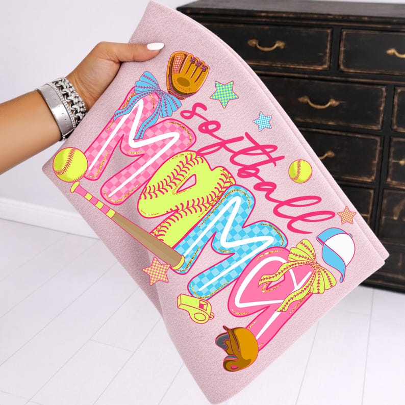 May include: A pink softball-themed towel with the word "MOM" in large, colorful letters. The design includes baseballs, a bat, glove, and stars. The word "softball" is written in cursive.