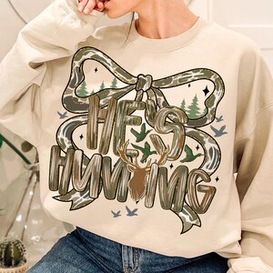 May include: A beige sweatshirt with the text "HE'S HUNTING" in a camouflage-style font, with a deer graphic. The design includes a bow, trees, and birds. The sweatshirt is a casual, comfortable style.