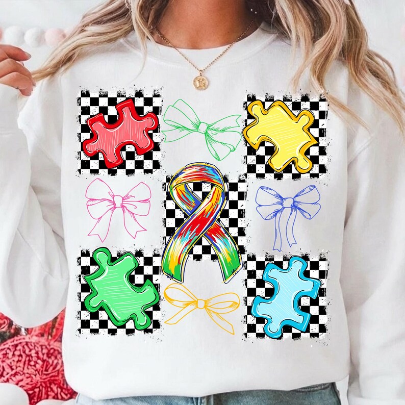 May include: White sweatshirt with a design of colourful puzzle pieces, bows, and a tie-dye ribbon. The graphics are set against a black and white chequered background. The design includes red, green, yellow, pink, blue, and yellow elements.