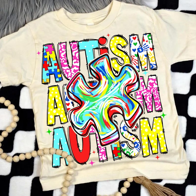May include: Cream-colored t-shirt with the word "AUTISM" in colorful, block letters. A large, multi-colored puzzle piece is centered on the shirt. The design includes hearts, handprints, and puzzle pieces.