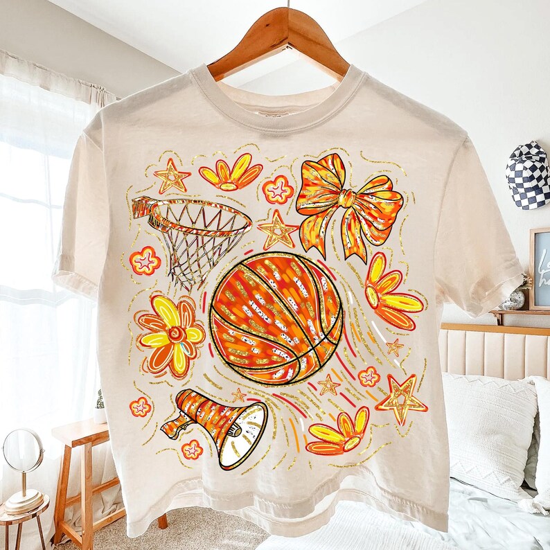 May include: Cream-colored t-shirt featuring a basketball design with a basketball, flowers, stars, and a bow. The basketball is orange with gold accents. The design includes a basketball hoop and a megaphone.
