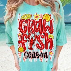 May include: A light turquoise t-shirt with a red and white graphic that says "Crawfish Season." The design includes a crawfish, lemons, and stars. The shirt has short sleeves and a relaxed fit.