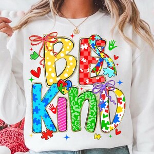May include: White sweatshirt with the words "BE KIND" in colorful, puzzle-piece-themed lettering. The design includes hearts, handprints, and bows, with a gold glitter outline. The letters are in various patterns and colors.