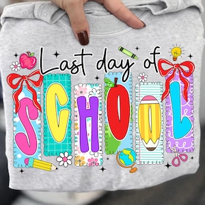 Puede incluir: Camiseta gris claro con un gráfico colorido que dice "Last day of SCHOOL". El diseño incluye una manzana, un lápiz, un globo terráqueo y unas tijeras. Las letras son de varios colores, con bordes decorativos y lazos.