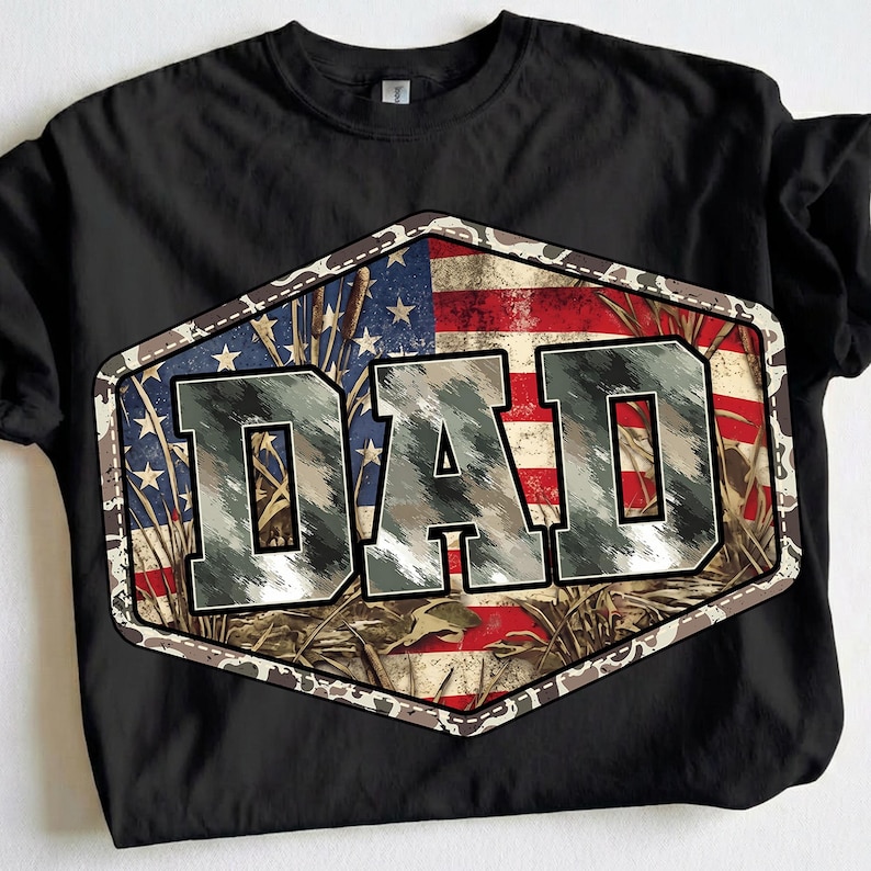 以下が含まれることがあります： 黒いTシャツ。迷彩柄で「DAD」の文字が描かれたグラフィックデザイン。アメリカ国旗と迷彩模様を組み合わせ、六角形のフレームで囲んでいます。