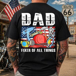 May include: Black t-shirt with the word "DAD" in white block letters above an American flag design. The flag graphic features tools like a hammer, wrench, and drill. The text "FIXER OF ALL THINGS" is below the flag.