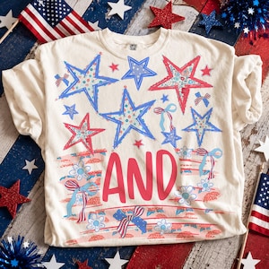 Puede incluir: Camiseta color crema con un diseño patriótico con estrellas rojas y azules, lazos y detalles florales. La palabra "AND" está impresa en rojo. La camiseta está decorada con un estilo vintage desgastado.